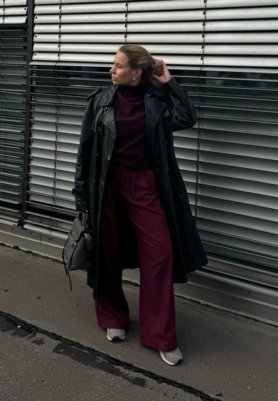 Abrigo de cuero negro, suéter de cuello alto burdeos, pantalones anchos color marrón oscuro, zapatillas grises, llevando una bolsa gris, de pie cerca de persianas horizontales.