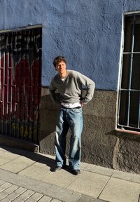 Pull gris clair à col en V, t-shirt blanc en dessous, jean en denim bleu ample, chaussures noires, debout contre un mur texturé bleu.