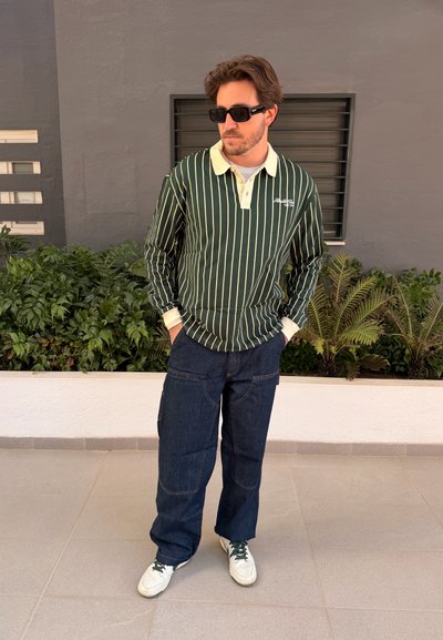 Hombre con gafas de sol negras, polo de manga larga de rayas verdes y blancas, vaqueros oscuros y zapatillas blancas, con las manos en los bolsillos.