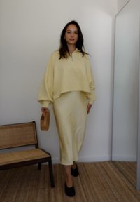 Ανοιχτό κίτρινο κοντό φούτερ με φερμουάρ στην κολλέτα, συνδυασμένο με μίντι φούστα στο ίδιο χρώμα. Το μοντέλο κρατάει ένα μικρό ξύλινο καλάθι. Καφέ πάγκοι στο παρασκήνιο.