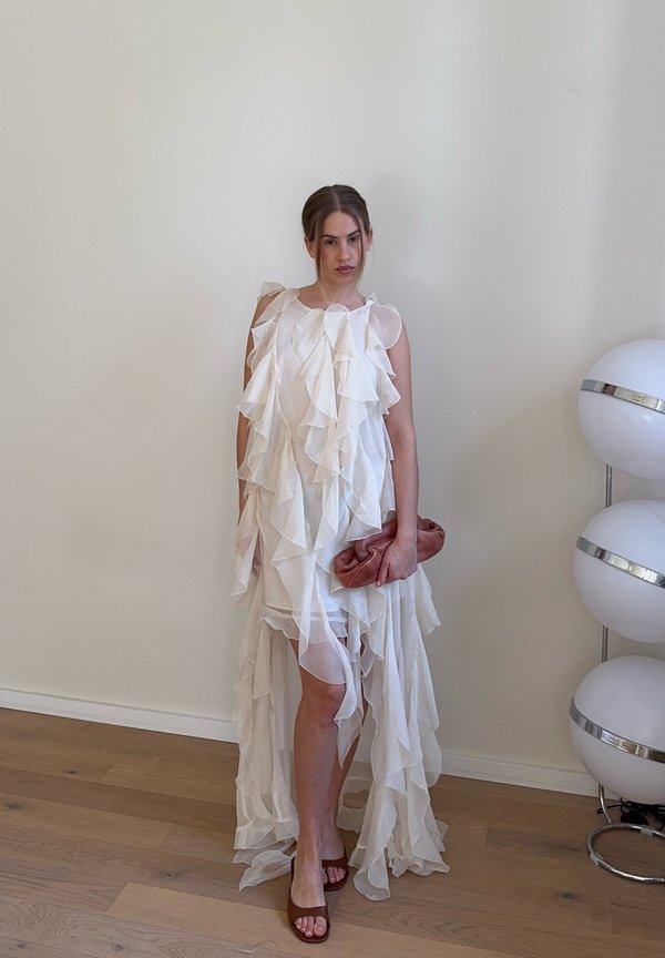 Frau in einem weißen, mehrlagigen Rüschenkleid, die eine rostfarbene Clutch hält und auf einem Holzboden neben einer weißen modernen Stehlampe steht.