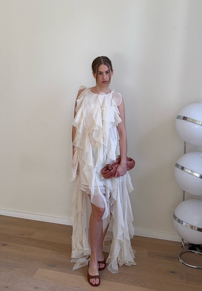 Ženska, oblečena v belo obleko z več plastmi volančkov, drži rjavkasto oranžno torbico, stoji na lesenem tlaku poleg bele moderne stoječe svetilke.