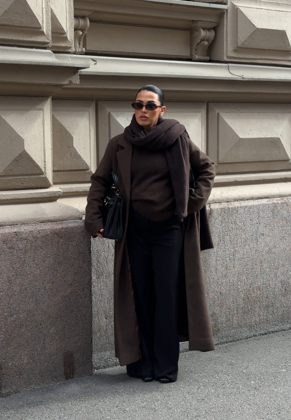 Palton lung maro peste un top închis la culoare și pantaloni largi; asortat cu o eșarfă și un geantă neagră; ochelarii de soare mici completează aspectul.