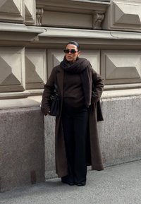 Palton lung maro peste un top închis la culoare și pantaloni largi; asortat cu o eșarfă și un geantă neagră; ochelarii de soare mici completează aspectul.