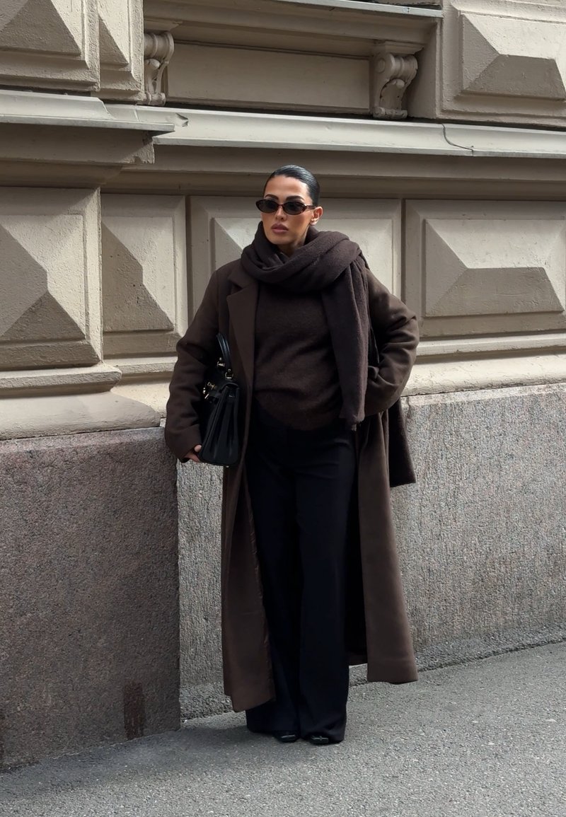 Bruine lange jas over een donker topje en wijde broekspijpen; gecombineerd met een bijpassende sjaal en een zwarte handtas; kleine zonnebril maakt de look af.