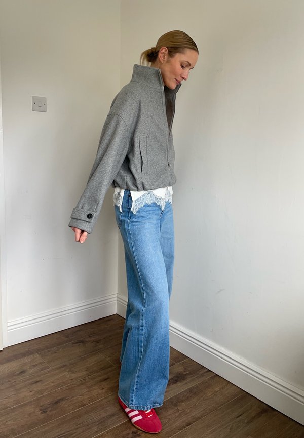 Kobieta stoi w pomieszczeniu, ubrana w szarą kurtkę na zamek, biały top z koronką, niebieskie dżinsy o szerokich nogawkach oraz czerwone trampki z białymi paskami.