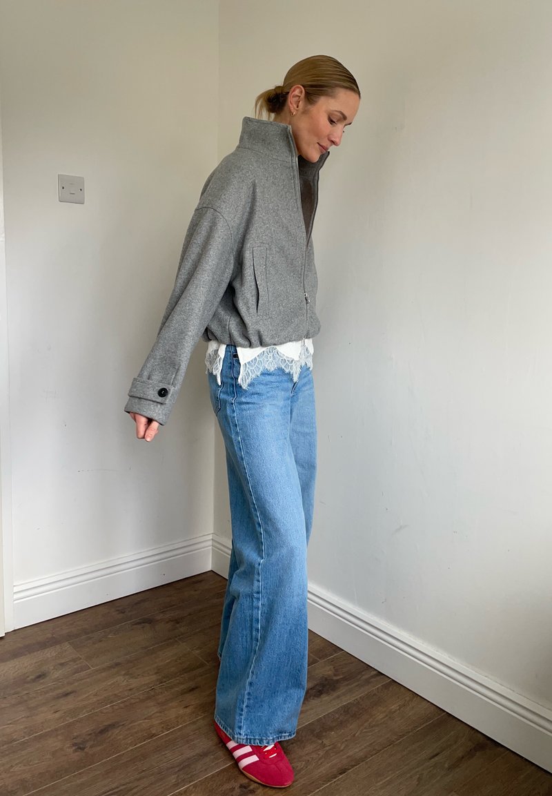 Femme debout à l'intérieur portant une veste grise à fermeture éclair, un haut blanc orné de dentelle, un jean bleu à jambes larges, et des baskets rouges à rayures blanches.