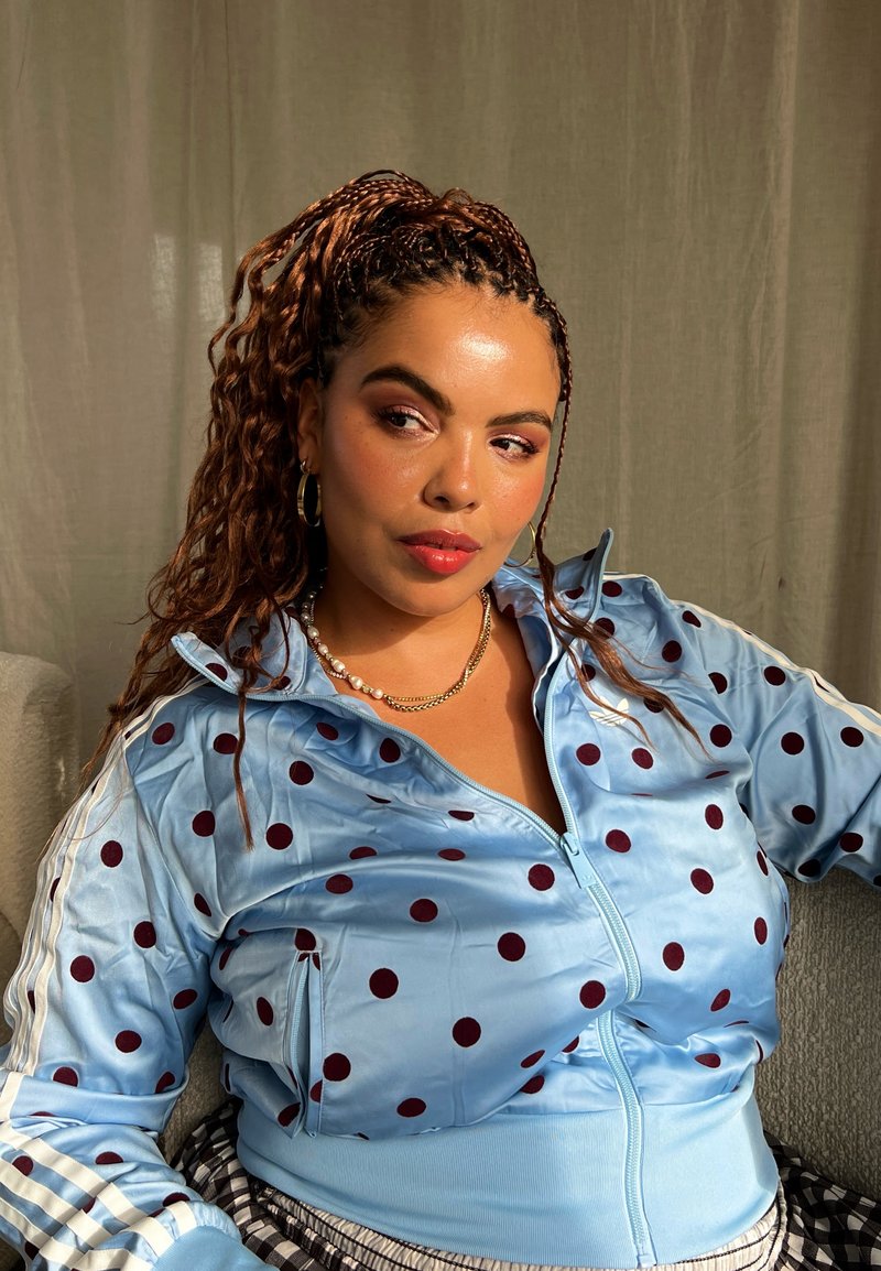 Une femme aux cheveux tressés est assise contre un rideau. Elle porte une veste bleu clair à pois bordeaux et plusieurs colliers dorés superposés.