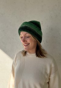 Gorro de malha verde às riscas, feito de lã suave, apresentando tonalidades alternadas de verde escuro e claro; usado com um suéter texturizado de cor creme.