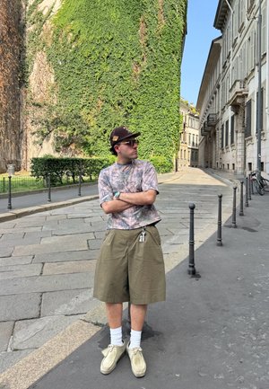 Camisa de manga corta con camuflaje, pantalones cortos verde oliva, calcetines blancos, zapatos beiges, gorra de béisbol, de pie en una calle empedrada cerca de vegetación.