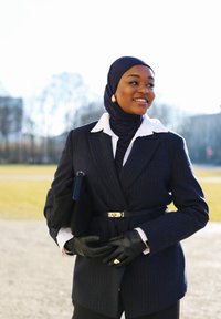 Femme portant un hijab noir, un blazer à fines rayures avec un col blanc, des gants en cuir noir et des boucles d'oreilles dorées, souriant en plein air en plein jour.
