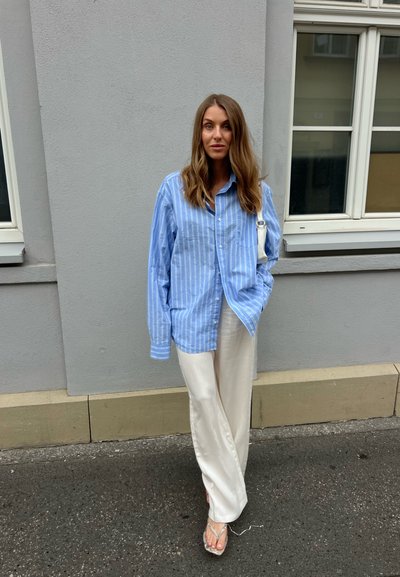 Mujer con cabello largo y castaño que lleva una camisa de rayas azul, pantalones blancos de pierna ancha y sandalias, de pie en una acera junto a un edificio gris.