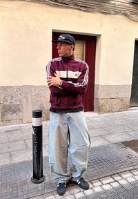 Jachetă zip-up maroon cu dungi albe și logo, blugi oversized de culoare albastru deschis, șapcă neagră și sneakers negri cu dungi albe, stând lângă un stâlp.