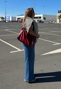Borsa a tracolla rossa in pelle, sciarpa a righe bianco e nera, jeans blu a gamba svasata, in piedi in un parcheggio sotto un cielo sereno e blu.