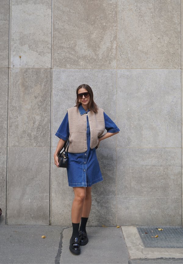 Femme portant des lunettes de soleil, un gilet beige sur une robe en denim bleue, des chaussettes noires et des chaussures noires, debout contre un mur en pierre gris.