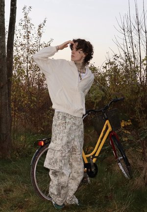 Persona con chaqueta blanca y pantalones estampados está de pie sobre césped junto a una bicicleta amarilla con cesta, mirando hacia la distancia.