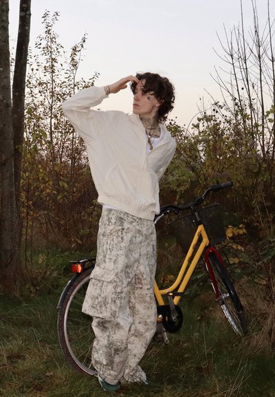 Persona con chaqueta blanca y pantalones estampados está de pie sobre césped junto a una bicicleta amarilla con cesta, mirando hacia la distancia.