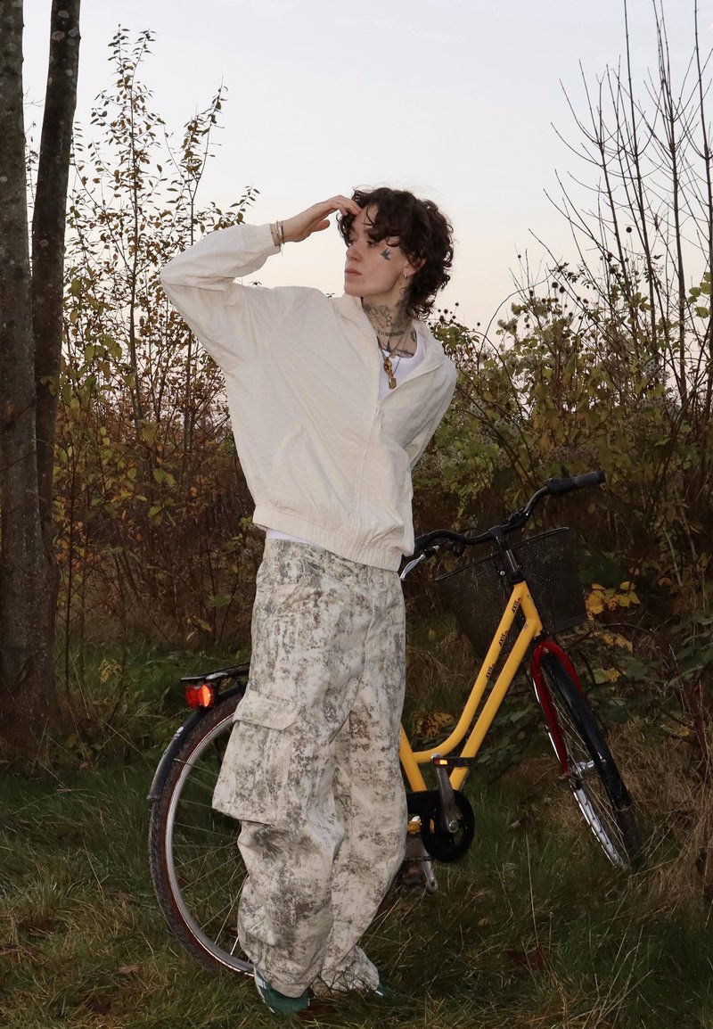 Persoană îmbrăcată într-o jachetă albă și pantaloni cu model stă pe iarbă lângă o bicicletă galbenă cu coș, privind spre depărtare.