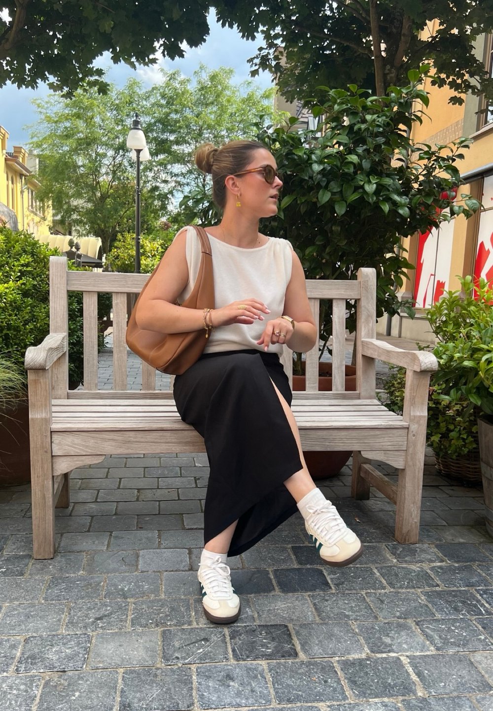 Μπεζ δερμάτινη τσάντα ώμου, κρεμ μπλούζα, μαύρη φούστα με πλευρική σχισμή και κρεμ αθλητικά παπούτσια με παστέλ λεπτομέριες, καθισμένη σε ξύλινο παγκάκι.