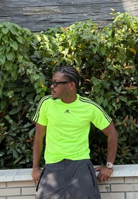 Neon rumena športna majica Adidas z črnimi črtami, v kombinaciji s sivo kratkimi hlačami. Oseba sedi ob ozadju listnate zelenja in kamnite stene.