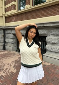 Gilet in maglia crema con scollatura a V e bordi verde scuro, abbinato a una gonna bianca a pieghe. Presenta una toppa con logo sul petto.