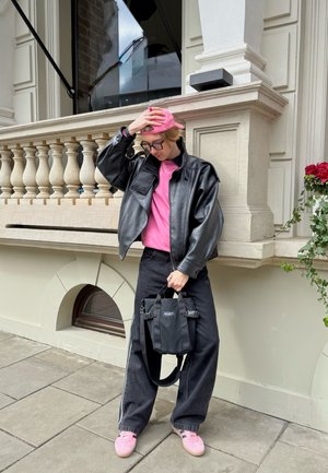 Persona que lleva una gorra rosa, camisa rosa, chaqueta de cuero negra y pantalones negros, sosteniendo una bolsa negra, de pie en una acera junto a un edificio de piedra.