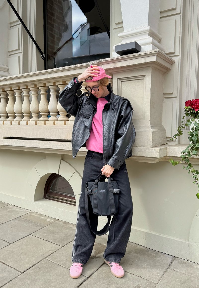 Személy rózsaszín sapkát, rózsaszín inget, fekete bőrkabátot és fekete nadrágot visel, fekete táskát fog, és egy járdán áll egy kőépület mellett.