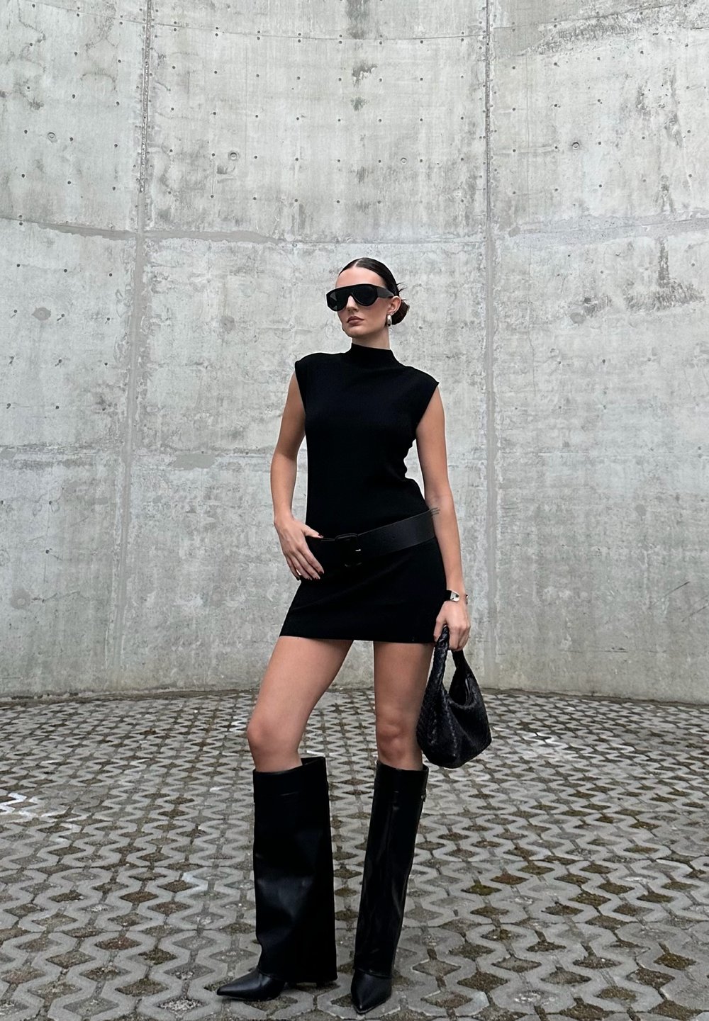 Zwart mouwloos jurkje met hoge kraag, gecombineerd met kniehoge zwarte leren laarzen en oversized zonnebril. Accessoires zijn een zwarte riem en handtas.