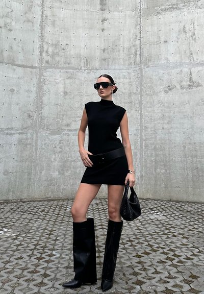Vestido negro sin mangas con cuello alto, combinado con botas de cuero negras hasta la rodilla y gafas de sol de gran tamaño. Los accesorios incluyen un cinturón negro y un bolso.