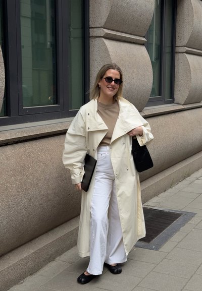 Mujer con gabardina larga crema, pantalones blancos y gafas de sol, sosteniendo bolsa negra y carpeta, de pie junto a un edificio de piedra en la acera.