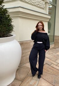 Suéter preto com decote à frente, calças largas pretas e sapatos pretos. Texturas suaves com um cinto branco como destaque. O fundo inclui um grande vaso branco.