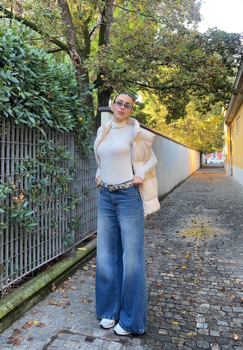 Chunky creme Puffer-Weste über einem taillierten beigen Rollkragenpullover, kombiniert mit weit geschnittenen blauen Jeans und weißen Sneakers. Sonnenbrille vervollständigt den Look.