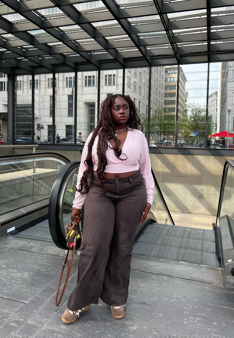 Femme aux longs cheveux, portant un haut rose et un pantalon foncé, se tient près d’un escalator dans une zone urbaine couverte de verre.