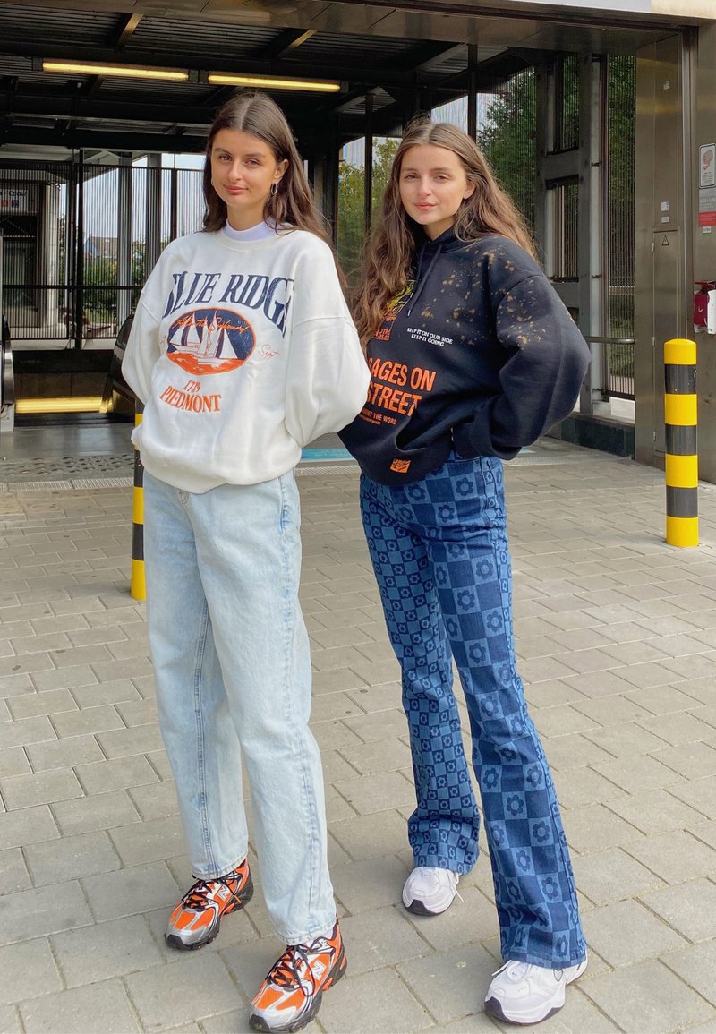 Hvid sweatshirt med grafisk design, lyseblå jeans, sneakers med orange detaljer. Sort sweatshirt med mønster, blå mønstrede flagrende jeans, hvide sneakers.