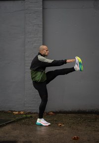 Scarpe da ginnastica atletiche con tomaia bianca e giallo neon, dettagli neri e verdi; persona che si allunga su uno sfondo grigio, indossando una giacca nera e verde.