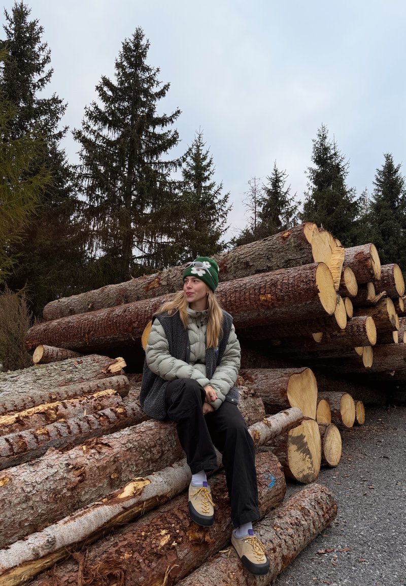 Veste grise rembourrée, gilet sombre, pantalon noir et chaussures marron. Assis sur des bûches empilées entourées d'arbres à feuilles persistantes sous un ciel nuageux.