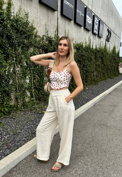 Top corto blanco con estampado de cerezas rojas, pantalones de lino beige anchos y sandalias deslizantes verdes. El fondo presenta una pared de concreto texturizada y hiedra.