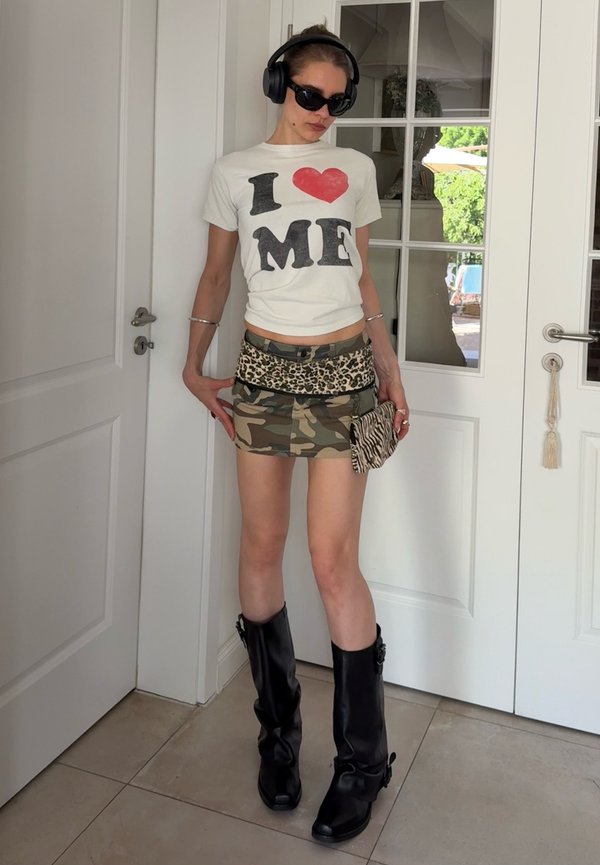 Frau trägt schwarze Kopfhörer, Sonnenbrille, weißes T-Shirt mit der Aufschrift "Ich liebe mich", Rock in Camouflage- und Leopardenmuster, schwarze Stiefel und hält eine Clutch mit Zebramuster.