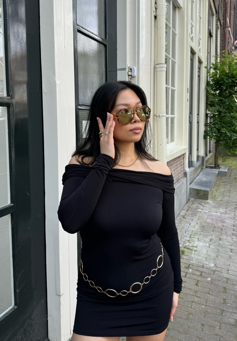 Vestito nero off-the-shoulder con maniche lunghe, dotato di una cintura in catena dorata. La modella indossa occhiali da sole grandi e riflettenti e gioielli.