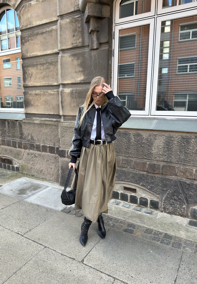 Femme aux longs cheveux blonds portant une veste en cuir noire, une chemise blanche, une cravate, une jupe midi beige, des bottes noires et portant un sac à main noir sur un trottoir en ville.