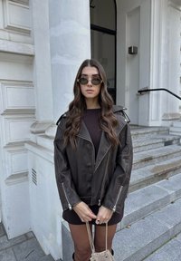 Smešasti kožni moto jakna s srebrnim patentnim zatvaračima, složena preko tamne haljine. Model drži torbu sa teksturom svjetle boje. Stepenice i bijeli stupovi u pozadini.