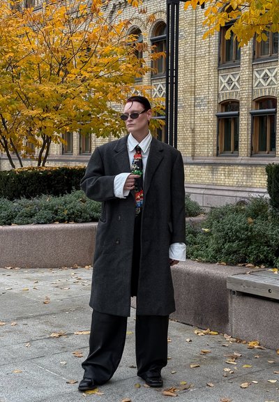 Abrigo de lana gris sobre conjunto negro, camisa blanca con puños destacados, corbata de colores con patrón, gafas de sol oscuras y zapatos negros. Follaje otoñal al fondo.
