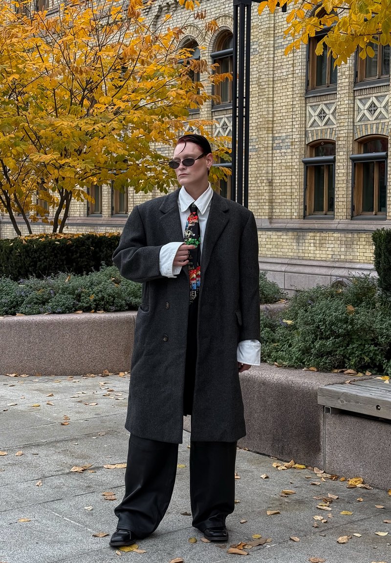 Manteau en laine grise sur une tenue noire, chemise blanche avec des poignets remarquables, cravate colorée à motifs, lunettes de soleil foncées et chaussures noires. Feuillage d'automne en arrière-plan.