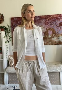 Chemise blanche à boutons avec deux poches poitrine, portée par-dessus un haut blanc court avec le logo "CK", associée à un pantalon beige à cordon de serrage.