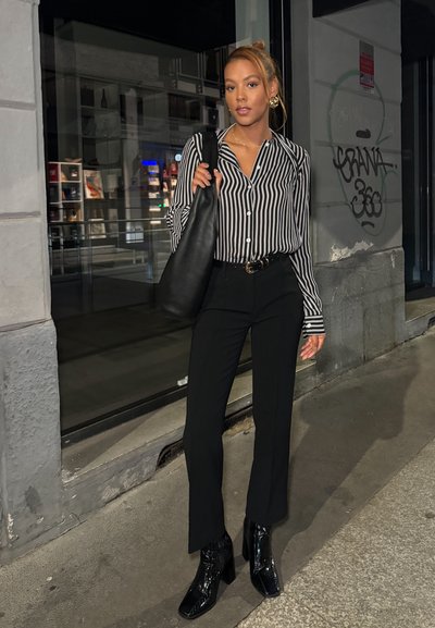 Camisa de botones a rayas blancas y negras, pantalones negros ajustados, botas de cuero negro de tobillo brillantes y un bolso de mano negro. Ambiente urbano nocturno.