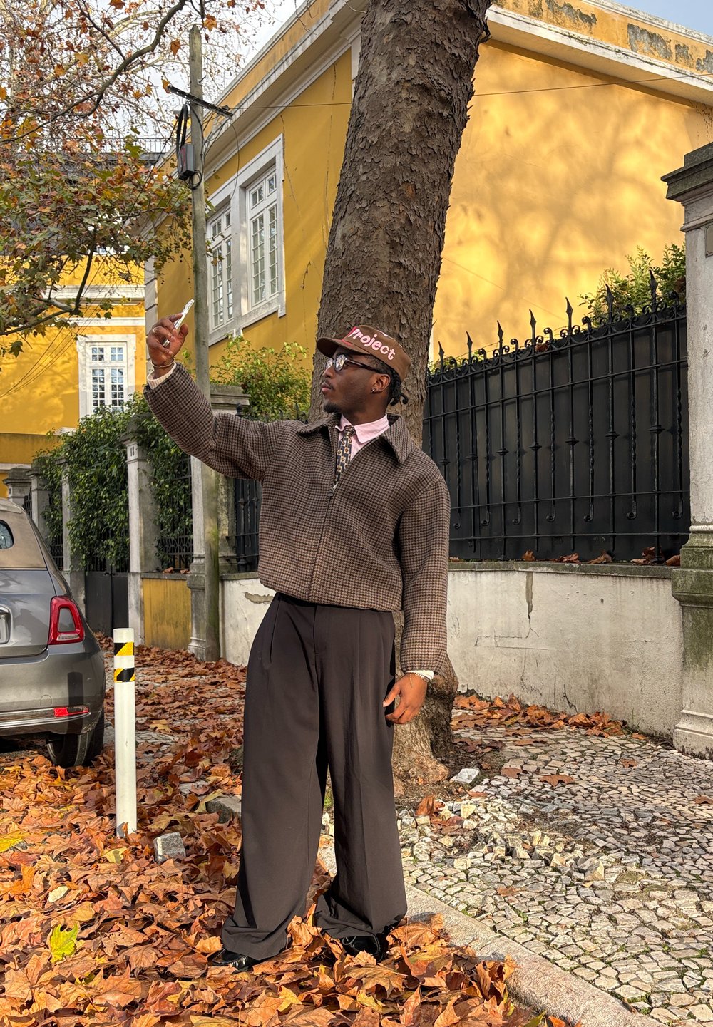 Veste à motif marron portée sur une chemise rose, associée à un pantalon large noir. Porte une casquette marron avec le texte « Project » et des lunettes, prenant un selfie.
