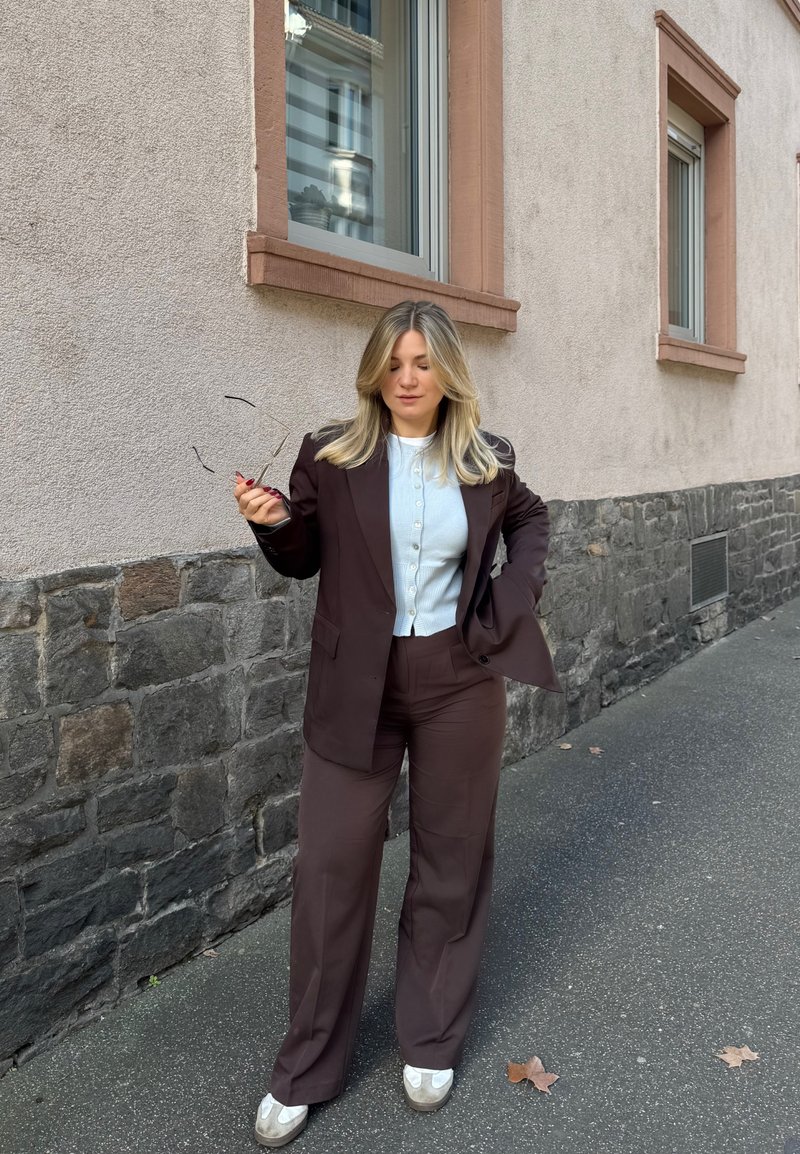 Femme aux cheveux blonds portant un costume marron et un haut blanc, debout sur le trottoir près d'un mur en pierre et plâtre, tenant des lunettes de soleil dans une main.