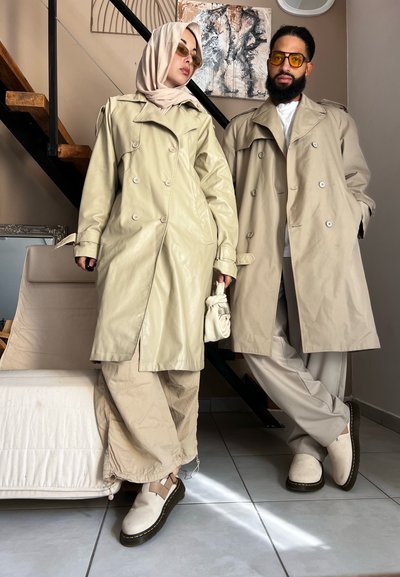 Abrigos de trench de piel beige de doble botonadura llevados por dos personas. Una de ellas lleva una bufanda ligera y texturizada, y ambas visten pantalones y zapatos en tonos neutros.