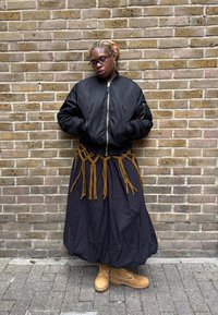 Sort bomberjakke over et flytende svart skjørt med brune frynser, kombinert med brune snøresko. Bakgrunn med murvegg.