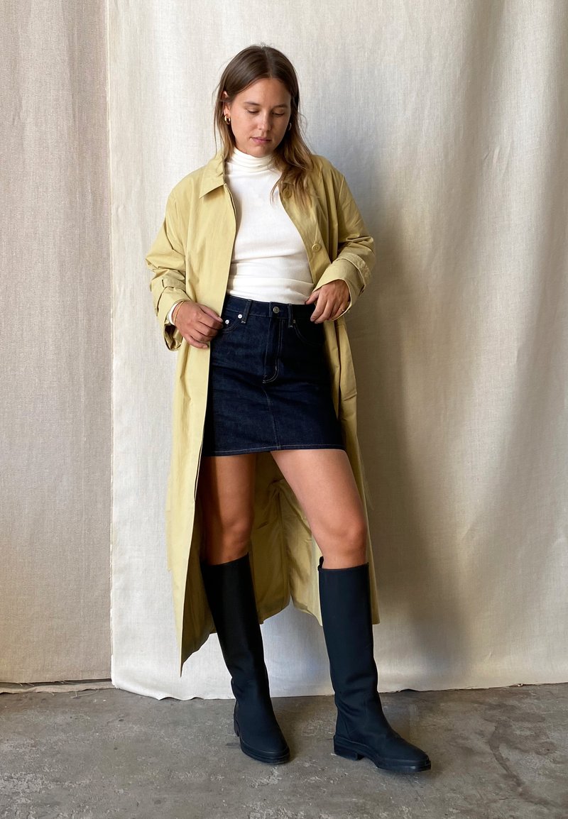 Pitkä vaaleankeltainen takki valkoisen korkeakaulaimen paidan päällä, yhdistettynä tummaan farkkuhameeseen ja polvimittaisiin mustiin saappaisiin. Sujuvat pinnat minimalistisella muotoilulla.